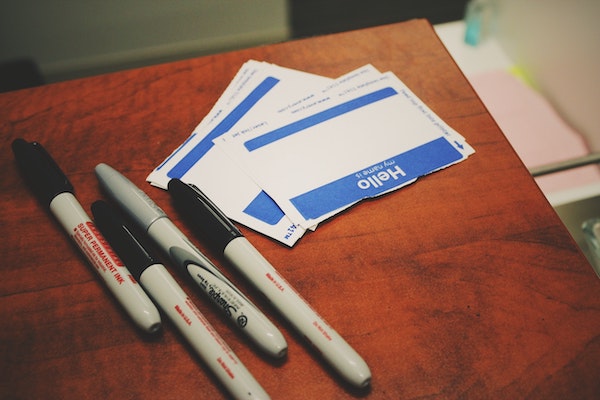 Blue "Hello My Name is..." stickerson a brown desk next to black sharpie pens.